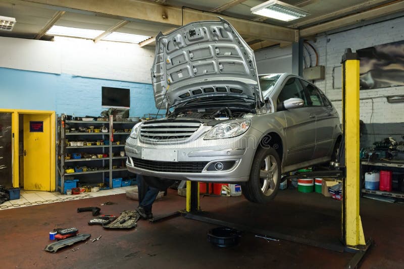 Silver car raised on a lift in a real auto repair garage with tools and shelves visible, showing a typical service visit that contributes to unexpected EV ownership costs