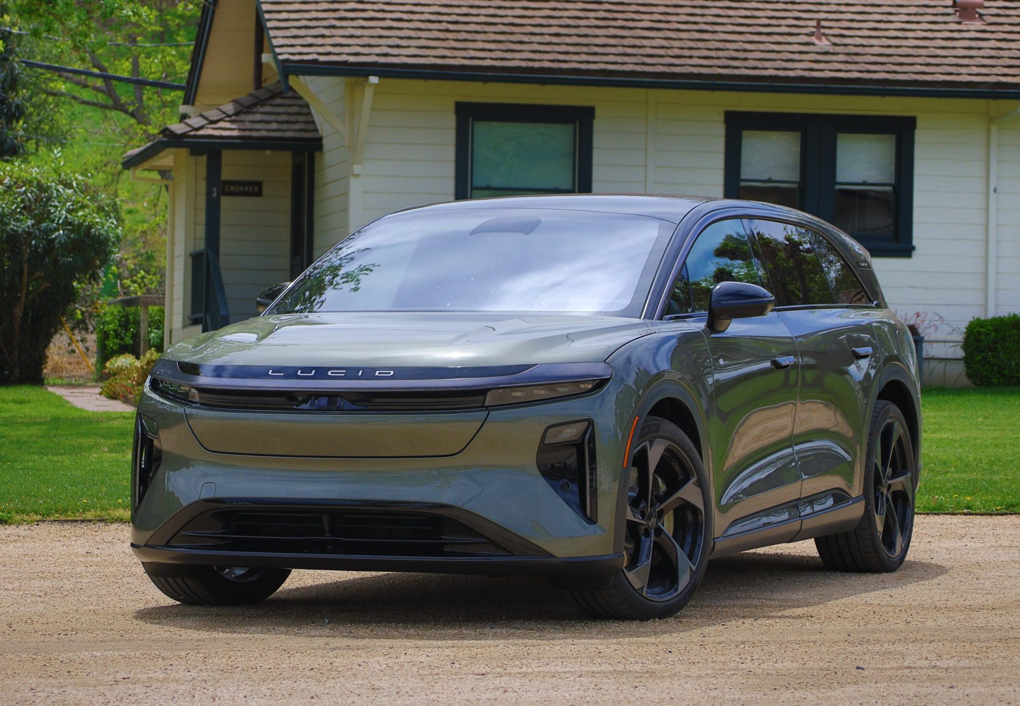 A premium dark green electric luxury SUV parked on gravel in front of a charming house with greenery, relaxed everyday luxury scene tied to anticipated electric vehicles launching in 2027