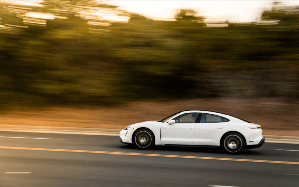 White Porsche Taycan driving on a scenic road with trees, representing luxury electric cars with good resale value retention after 3 years