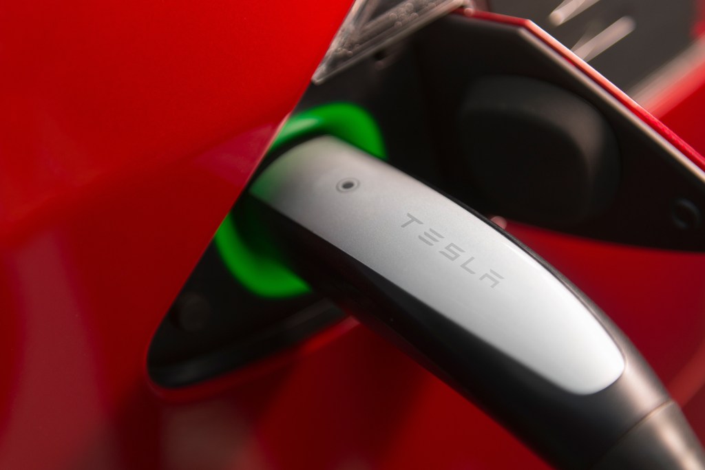 Real photo of a Tesla NACS charging connector plugged into the port of a red electric car, highlighting the compact design of the North American Charging Standard