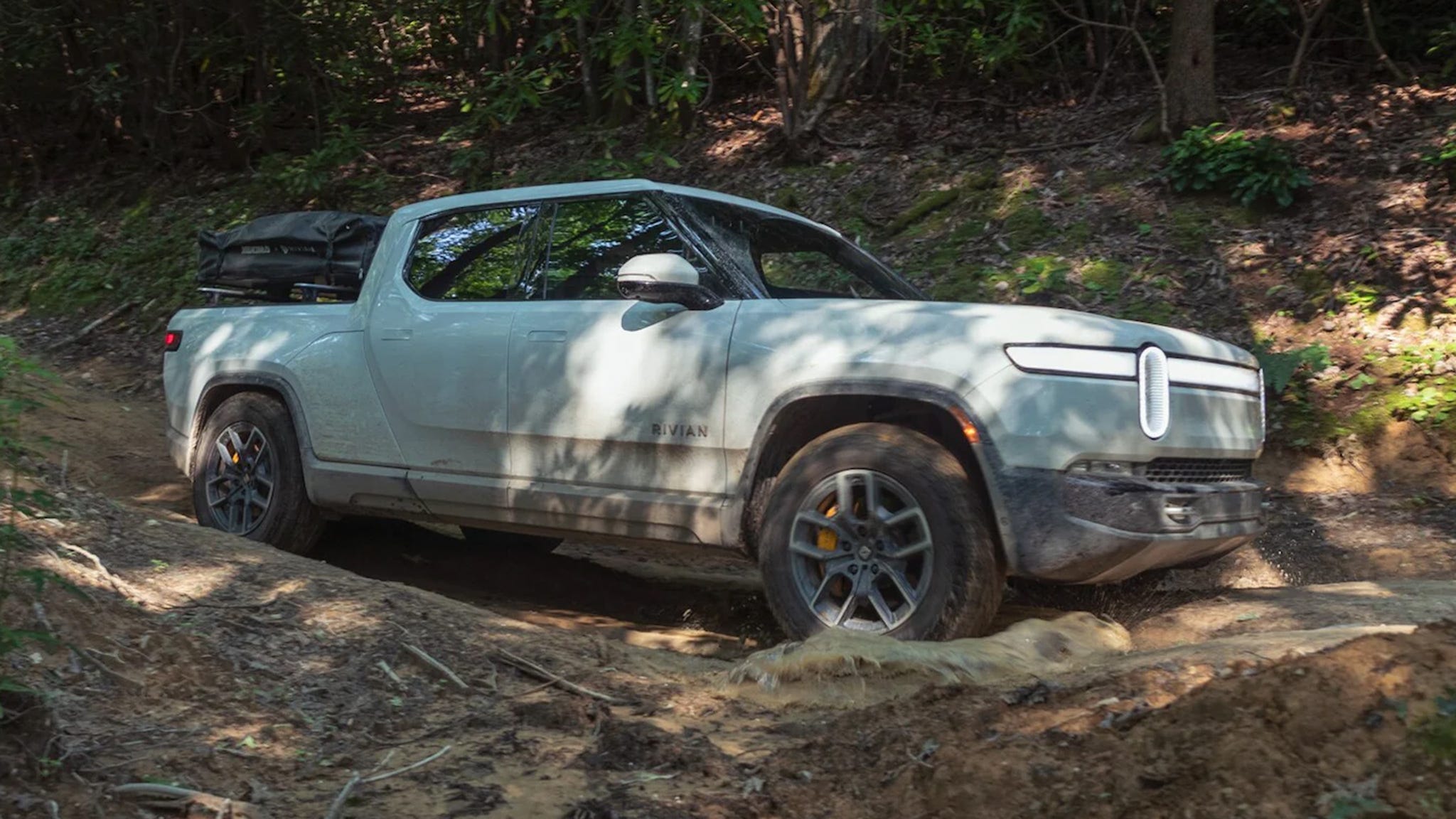 Rivian R1T tackling a muddy forest trail with gear on the roof, highlighting its adventure-ready capabilities among electric pickup trucks