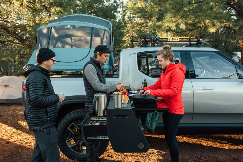 People cooking outdoors next to their electric truck at a forested campsite, showing how portable EV chargers enable charging while enjoying nature on road trips