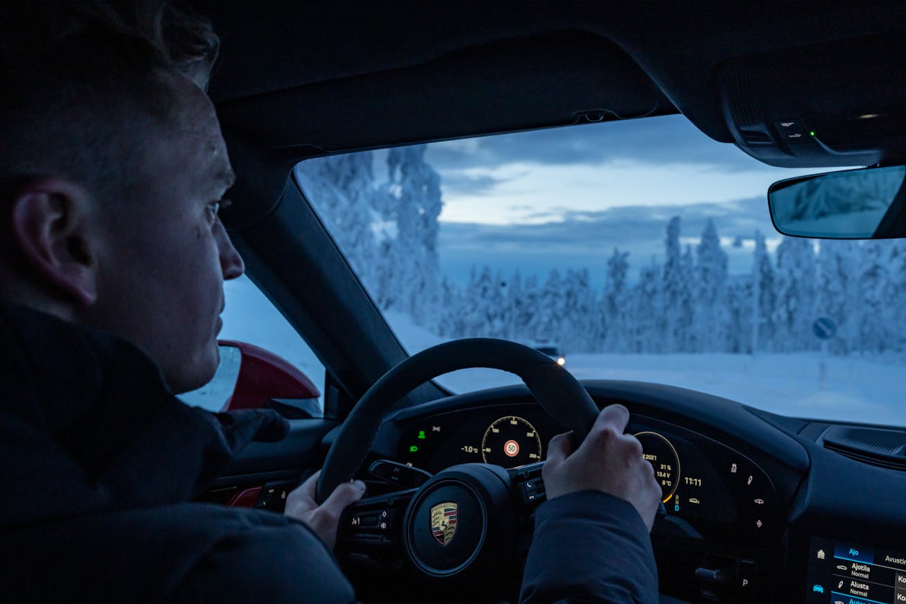 Driver inside an electric vehicle on a snowy winter road with frosted trees, enjoying improved range and comfort from preconditioned battery and warm cabin
