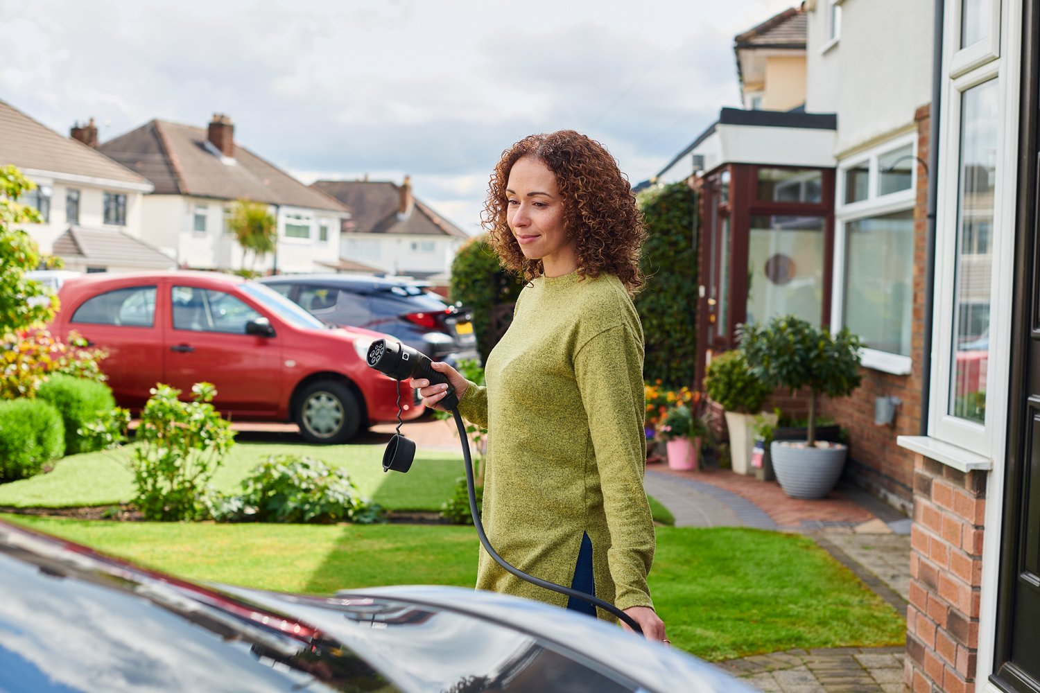 Person plugging in their electric vehicle at home driveway in the evening, relaxed moment with home charger like Tesla Wall Connector or JuiceBox