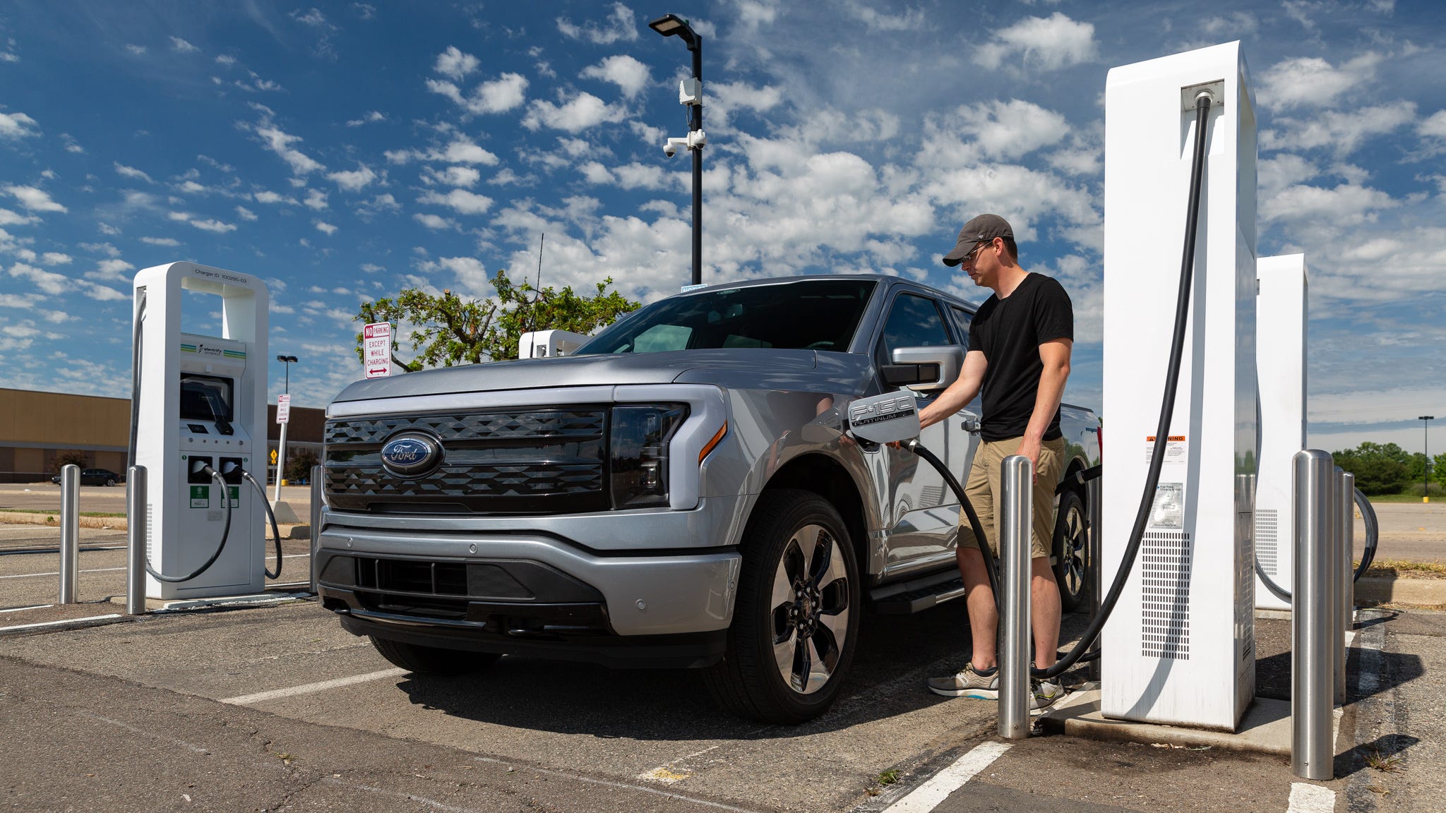Person plugging in a gray Ford F-150 Lightning at a public charging station on a sunny day, capturing the practical reality of owning an electric pickup truck