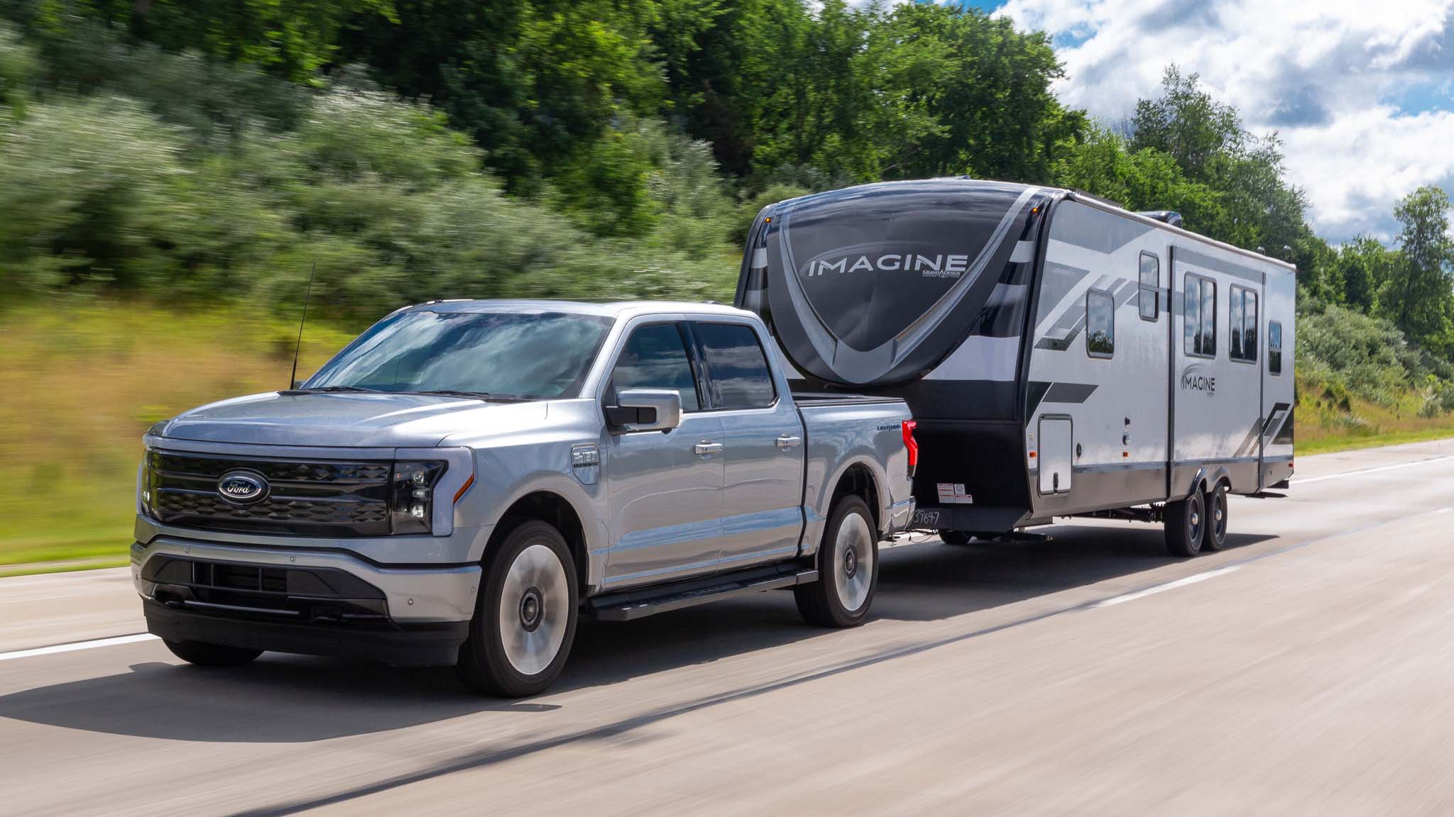 Ford F-150 Lightning towing a travel trailer on a highway surrounded by green trees, demonstrating real towing capability of electric pickup trucks