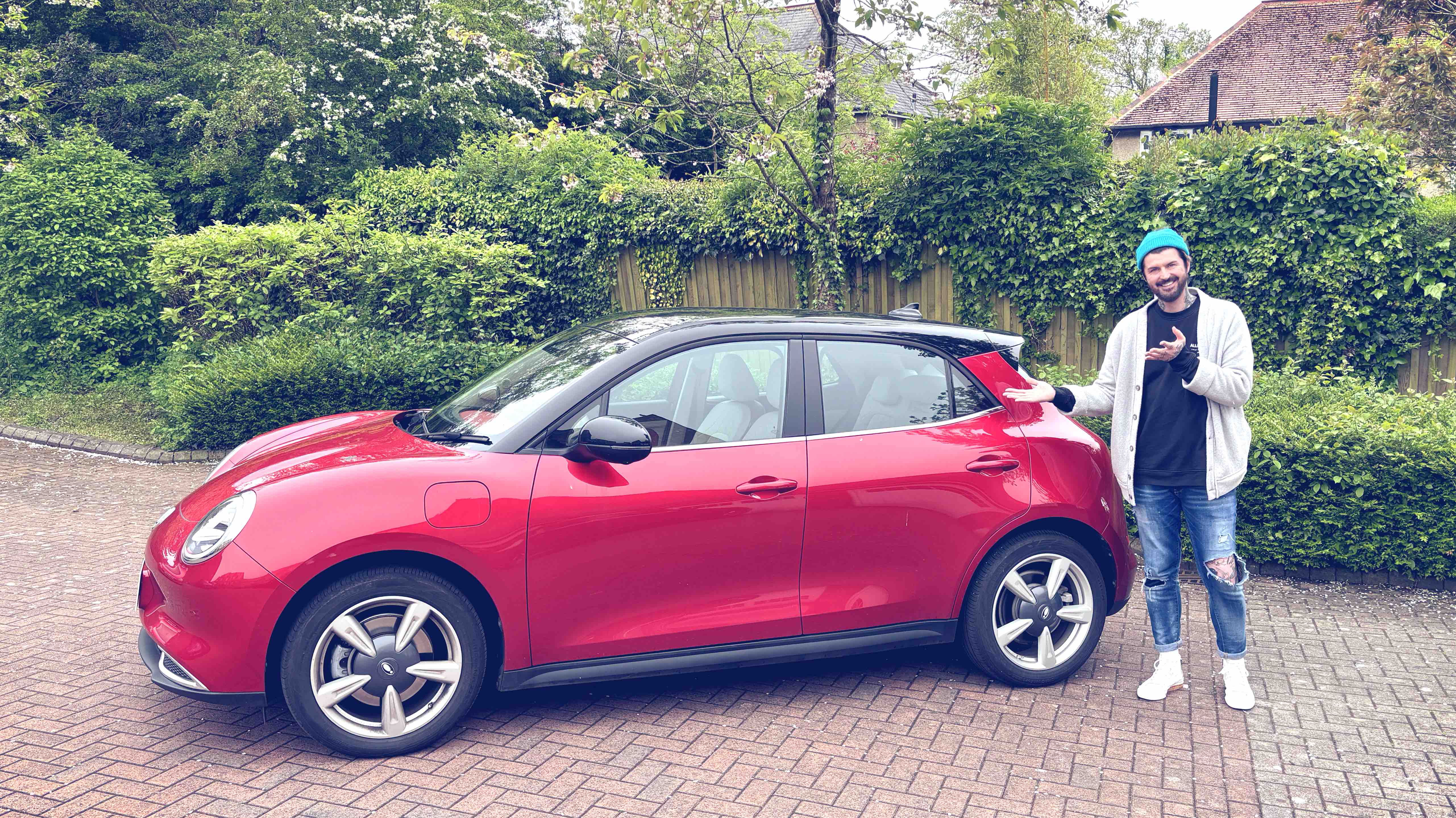 A young person stands next to a compact red electric hatchback parked in a residential driveway surrounded by greenery, everyday real-life scene that reflects accessible upcoming electric vehicles in 2027