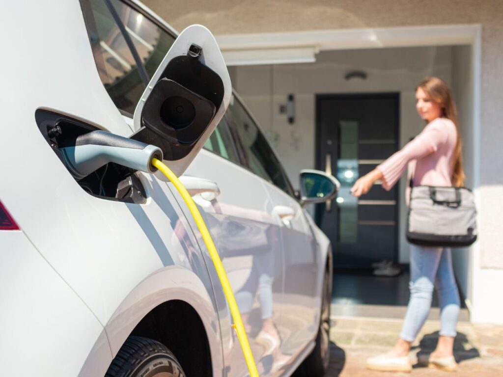 Diverse woman casually plugging the charger into her white electric sedan outside a modern home garage, everyday home EV charging moment with natural light