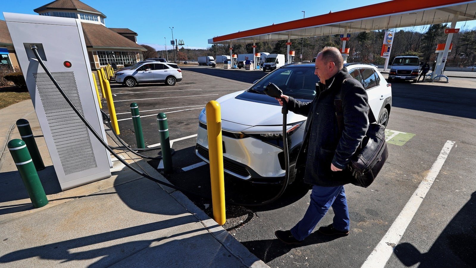 A driver plugging in a white electric SUV at a public charging station in a shopping area parking lot, highlighting free EV charging opportunities at malls and stores