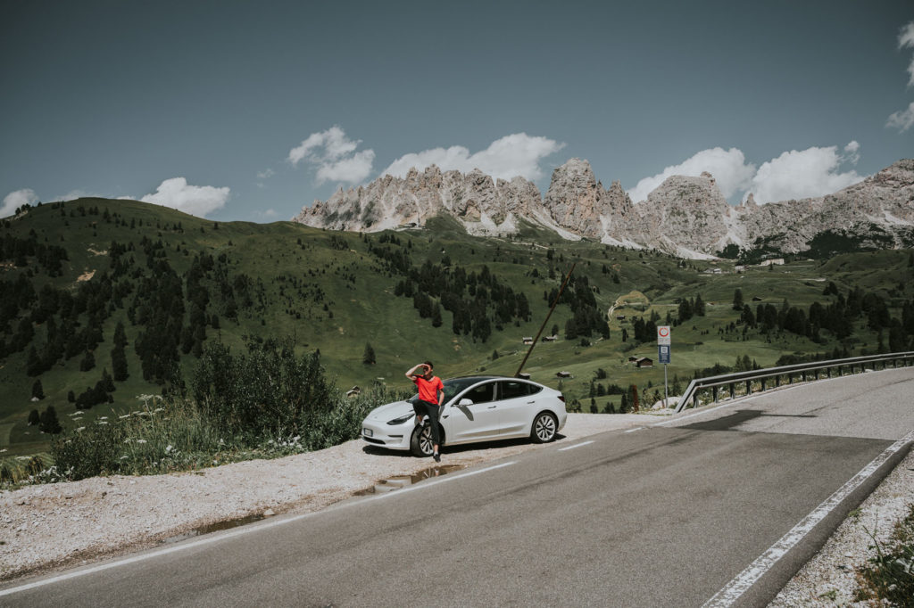 Tesla driver relaxing against their white Model 3 parked on a scenic mountain road with dramatic peaks in the background, evoking the freedom of EV road trips powered by supercharging