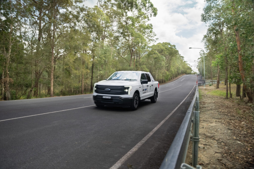 White Ford F-150 Lightning cruising down a tree-lined rural road on a sunny day, showing the practical side of electric pickup trucks in everyday driving