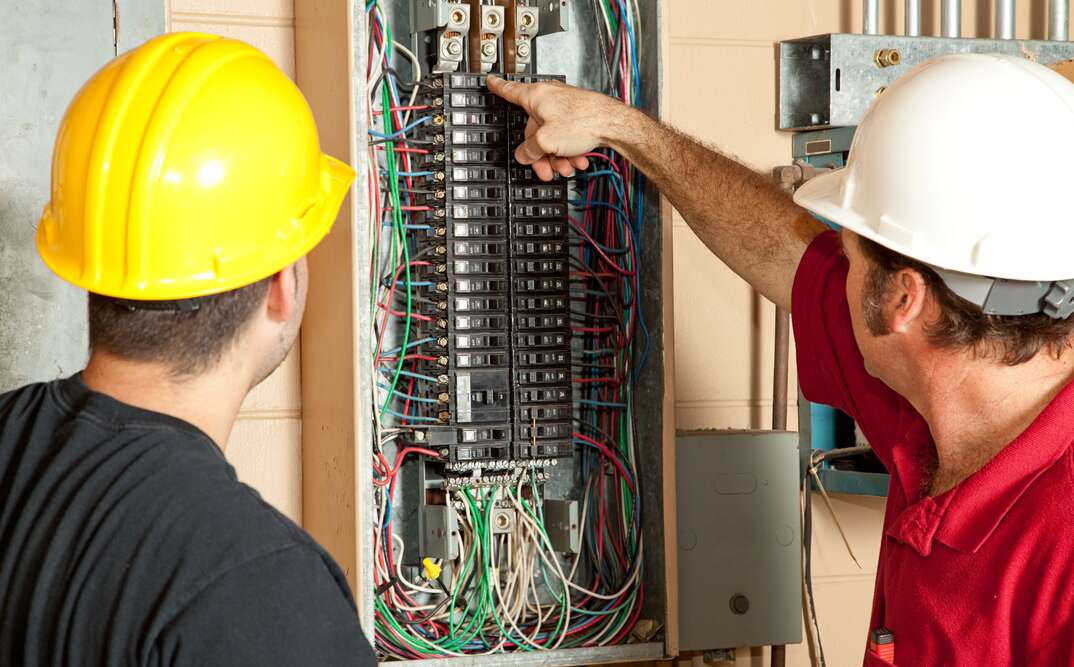 Two electricians examining an open home electrical panel with visible wires and breakers, showing the complexity involved in preparing for safe EV charger installation at home